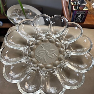 May include: A clear glass egg serving platter featuring a scalloped edge with twelve individual oval indentations for eggs. The center of the platter displays a frosted, embossed design of a poinsettia flower with leaves and berries.