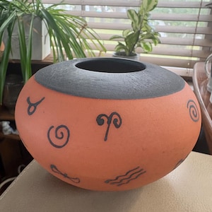 May include: A round, terracotta-coloured ceramic bowl with a wide, dark grey textured rim. The exterior surface features various black hand-drawn symbols, including spirals, wavy lines, and zodiac-style icons, distributed across the orange body.