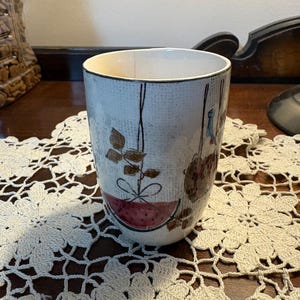 Pode incluir: Uma caneca de cerâmica com fundo azul claro e um design decorativo com uma melancia, folhas e cestos suspensos. A caneca tem interior branco e uma borda escura. Está sobre uma toalha de crochê creme.