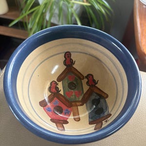 May include: A ceramic bowl with a tan interior and a dark blue rim. The interior features a hand-painted design of three birdhouses in red, green, and blue, each topped with a small red bird. The green birdhouse is decorated with red hearts.