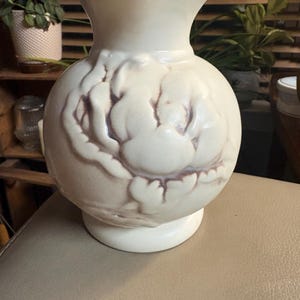 May include: A cream-colored ceramic vase with a rounded body and a flared rim. The exterior features a raised, embossed floral design with subtle brown shading in the crevices of the petals, set against a smooth, matte-finished surface.