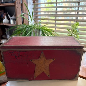 May include: A rectangular wooden box with a distressed red painted finish featuring a prominent dark brown metal star centred on the front panel. The box has a flat lid and visible cracks in the red paint, showing dark wood underneath.