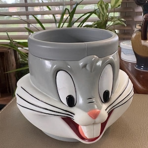 Pode incluir: Uma caneca de cerâmica cinza em forma de cabeça do Pernalonga, apresentando grandes olhos brancos com pupilas pretas, um pequeno nariz rosa, dentes frontais brancos proeminentes e bigodes pretos pintados em ambos os lados do rosto.