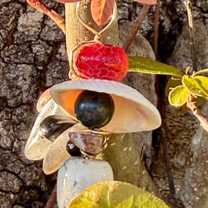 Może przedstawiać: Ręcznie robiony naszyjnik z wisiorkiem z czerwoną, teksturowaną perłą, białą muszlą i różnymi kamieniami w pionowym układzie. Wisiorek zawieszony jest na złocistym drucie i wisi na tle kory drzewa i liści.