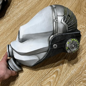 Puede incluir: Un casco blanco y plateado con una base negra, con un diseño detallado. El casco tiene una forma redondeada con una visera y un aparato respiratorio. El lateral del casco tiene un diseño circular con detalles verdes. El casco es probablemente un accesorio o una pieza de vestuario.