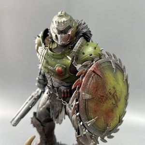 May include: A detailed figurine of a character in green and silver armour, holding a shield and a weapon. The shield is circular with a saw-blade edge and a blood-splatter design. The figure has a helmet and a fur-like collar.