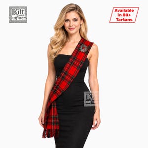 Peut inclure: Une écharpe en tartan à carreaux rouges avec des rayures noires et jaunes, ornée d'une broche circulaire argentée avec une pierre rouge centrale. L'écharpe a des extrémités frangées et est portée sur une robe noire. Le texte indique Kilt Outfitters et Available in 80+ Tartans.