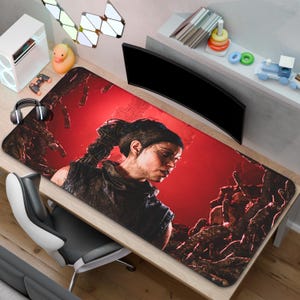 May include: A large rectangular gaming mousepad featuring a woman with a braid against a red background. The mousepad has a detailed graphic design and is placed on a wooden desk. Other items on the desk include a computer monitor, headphones, and a gaming console.