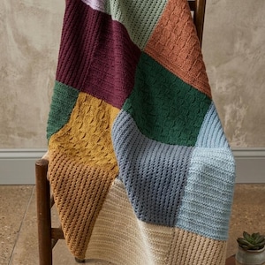 May include: A colorful, patchwork knitted blanket draped over a wooden chair. The blanket features various textured squares in shades of purple, green, orange, blue, and tan. The chair is a simple, vintage design.