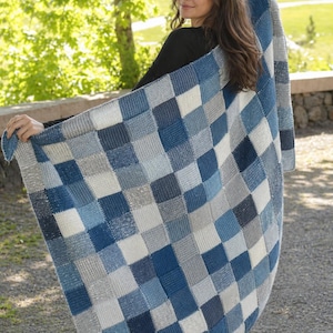 Peut inclure: Une couverture tricotée à carreaux bleus et gris. La couverture présente un motif de carrés dans différentes nuances de bleu et de gris. La couverture est tenue par une personne en extérieur.