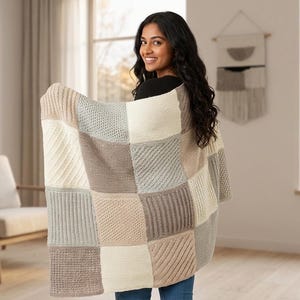 Könnte beinhalten: Eine rechteckige Strickdecke mit einem Patchwork-Design aus abwechselnden Quadraten in den Farben Creme, Beige, Taupe und Hellgrau. Die Quadrate weisen verschiedene Texturen auf, darunter diagonale Rippenmuster und vertikale Rippen.