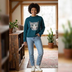 May include: Teal-colored sweater featuring a cat face design. The cat's face is gray and white with black detailing. The sweater is paired with light blue jeans and white sneakers. The woman is standing in front of a wooden bookshelf.