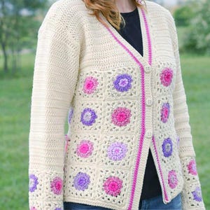 Op de afbeelding: Crèmekleurig gehaakt vest met een V-hals en knoopsluiting. De trui heeft vierkante bloempatronen in roze en paars, met roze bies. De mouwen hebben ook bloemaccenten.