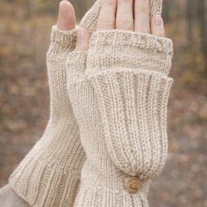 May include: Beige knitted fingerless gloves with a button fastening. The gloves are made from a thick, textured yarn and have a ribbed cuff. The mitten cover folds over the fingers and is secured with a wooden button.