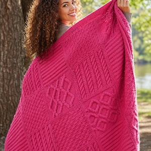 May include: A bright pink knitted blanket featuring a patchwork of textured patterns, including cable and geometric designs. The blanket is held up, displaying its intricate details and rich colour. The blanket appears soft and cosy.