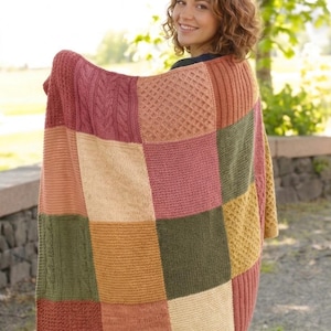 Puede incluir: Una manta de retazos multicolor en tonos rosa, verde, beige y naranja. La manta presenta varios patrones de tejido texturizados, incluidos trenzados y patrones de diamantes. La manta está siendo sostenida por una persona.