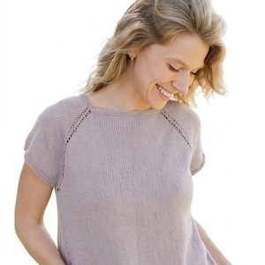 May include: A light purple knit top with short sleeves. The top has a square neckline and a textured knit pattern. The shoulders have a decorative open knit design. The model is smiling and looking down.
