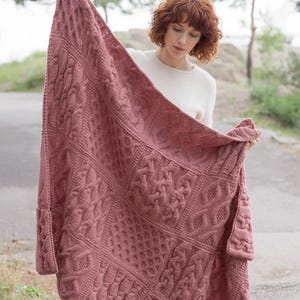 Può includere: Una grande coperta lavorata a maglia color rosa antico con intricati motivi a treccia e punti strutturati. La coperta rettangolare, con un bordo, viene tenuta in alto per mostrare il suo design. Il tema è l'arredamento accogliente della casa.