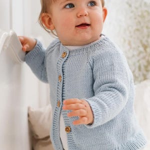 May include: A light blue long-sleeve knit cardigan featuring a round neckline and a front button closure with five small, round, light brown wooden buttons. The garment is constructed with a ribbed texture along the cuffs, hem, and button placket.