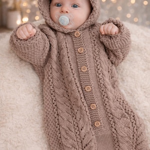 May include: A baby wearing a hooded, button-down, knitted jumpsuit in a soft brown color. The jumpsuit features a cable knit design and wooden buttons. The baby has blue eyes and a pacifier.