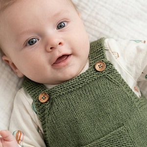 Könnte beinhalten: Ein olivgrüner, handgestrickter Baby-Overall mit zwei Holzknöpfen an den Schulterträgern, getragen über einem cremefarbenen Langarmshirt mit Heißluftballon-Muster. Der Text "HAND-KNITTED COLLECTION" ist in der unteren Ecke zu sehen.
