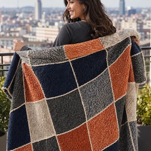 May include: A patterned blanket in navy blue, orange, grey, and beige, with a geometric design. The blanket is made of a textured material and features a grid-like pattern. The blanket is being held up, showing its size and design.