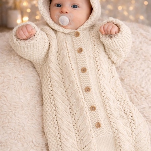May include: A baby in a cream-coloured, hooded knitted babygrow with a cable knit pattern and wooden buttons. The baby has a dummy and is lying on a fluffy, cream-coloured blanket.