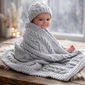Könnte beinhalten: Ein Baby, eingehüllt in eine graue Zopfstrickdecke und einen passenden Hut. Die Decke hat ein strukturiertes Design und ist über dem Baby drapiert, das auf einer Holzoberfläche sitzt. Das Baby lächelt und schaut in die Kamera.