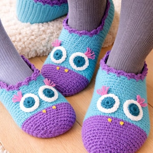 Peut inclure: Chausson en crochet faits à la main, bleus et violets, sur le thème du hibou. Les chaussons présentent des yeux blancs et bleus, des accents de plumes roses et un bec jaune. Bordure violette festonnée. Portés avec des chaussettes grises.