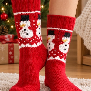 May include: A pair of red knitted socks featuring a white band with a repeating pattern of snowmen wearing black top hats and orange carrot noses. The socks have a ribbed cuff at the top and a solid red foot section, crafted with a thick, textured yarn.