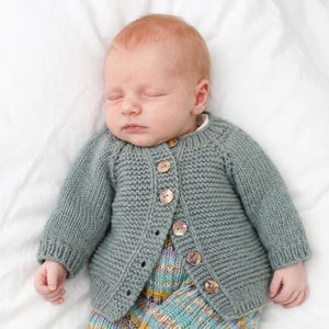 May include: A sage green, long-sleeved, hand-knitted cardigan featuring a round neckline and a front button closure with six round, mottled brown buttons. The garment is crafted with a textured garter stitch pattern and ribbed cuffs.