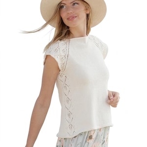 May include: A cream-coloured knitted top with short sleeves and a round neckline. The top features a decorative openwork design along the sleeves and sides. The person is wearing a wide-brimmed straw hat and a floral skirt.