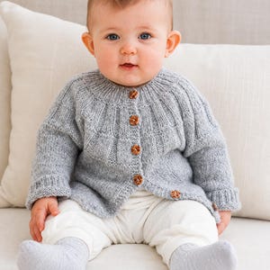 Puede incluir: Una chaqueta de punto gris claro con canesú acanalado y cinco pequeños botones marrones moteados en la parte delantera. La prenda de manga larga tiene cuello redondo y puños acanalados, ofreciendo un aspecto texturizado y uniforme.