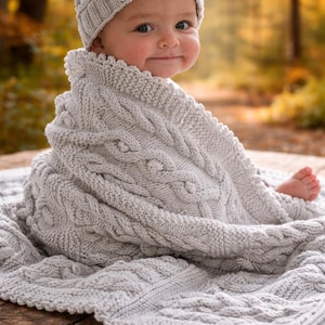May include: A baby wearing a light gray knitted hat and wrapped in a matching cable-knit blanket. The blanket has a textured design and is draped over a wooden surface. The background is blurred with autumn colors.
