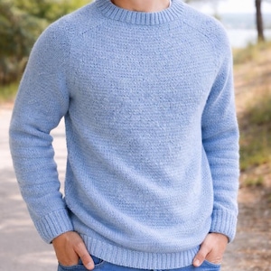May include: A light blue knit sweater with a crew neck and long sleeves. The sweater has a textured appearance and ribbed cuffs and hem. The person is wearing blue jeans. The background is a blurred outdoor scene.