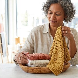 May include: A stack of three folded, textured knit cloths in mustard yellow, terracotta, and cream, resting in an oval wooden bowl. The mustard yellow cloth features a geometric pattern and is held upright, while the others remain stacked.