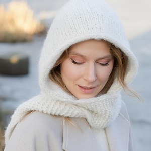 Può includere: Un passamontagna lavorato a maglia color crema con un motivo a coste testurizzato. Il copricapo presenta una parte superiore arrotondata e un'ampia sezione per il collo che ricade sulle spalle, realizzato in filato spesso e morbido.