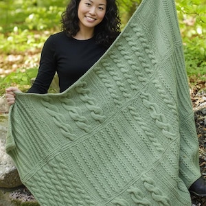May include: A sage green knitted blanket with a variety of cable knit and ribbed patterns. The blanket is displayed, highlighting its texture and design. The blanket looks soft and cosy, suitable for home decor.