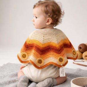 May include: A baby wearing a handmade crochet cape in cream, orange, and yellow chevron stripes. The cape has three large, round, yellow buttons and a scalloped edge. The baby is wearing a white onesie and gray socks.