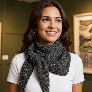May include: A dark grey knitted scarf with gold and white flecks is worn around the neck. The scarf is knotted at the front and worn over a white t-shirt. The woman in the image is smiling and looking to the side.