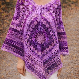 Könnte beinhalten: Handgefertigter, violetter Häkelponcho mit V-Ausschnitt und Kordelzug. Der Poncho hat ein geometrisches Muster mit verschiedenen Lilatönen, das ein strukturiertes Design erzeugt. Die Ärmel sind kurz und der Poncho hat eine bequeme Passform.