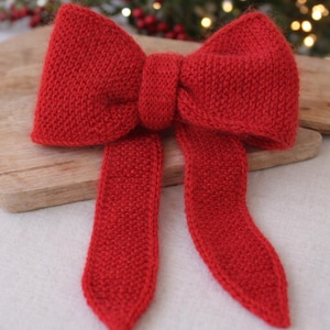 May include: A large, knitted red bow with a textured appearance. The bow features a central knot and long, flowing tails. The bow is likely a decorative item, possibly for the holidays or gift wrapping.