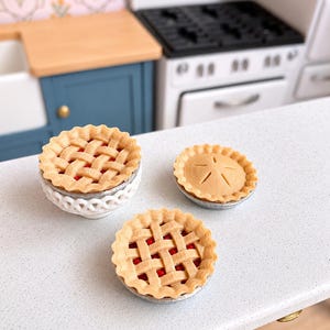 May include: Three miniature pies in a dollhouse kitchen setting. Two pies feature a golden-brown lattice crust revealing a red berry filling, while the third smaller pie has a solid crust with a star-shaped vent. The pies are set on a counter.
