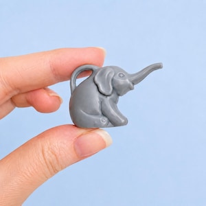 May include: A small, solid gray elephant figurine depicted in a seated position. The elephant features a smooth, matte surface, rounded ears, and an elongated trunk that extends upward and outward, showcasing a simple, molded design.