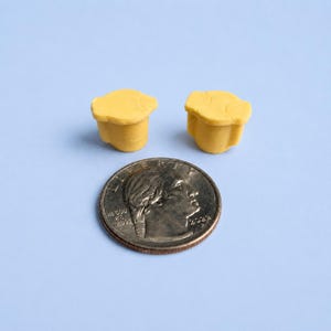 May include: Two small, yellow, three-dimensional objects with a rounded, irregular top and a cylindrical base. The objects are positioned above a standard United States quarter coin, which serves as a scale reference for their compact size.