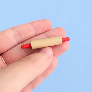 May include: A miniature rolling pin featuring a smooth, light tan cylindrical barrel and bright red handles on each end. The handles have a ribbed texture, and the central barrel is plain and unadorned, designed for small-scale crafting or display.