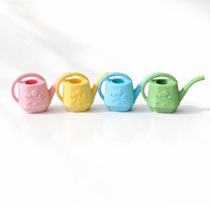 May include: Four small, pastel-colored watering cans. The colors are pink, yellow, blue, and green. Each can has a handle, a spout, and a floral design on the front. These miniature watering cans are ideal for small plants or decorative purposes.