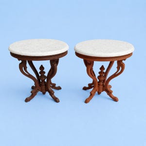 May include: Two miniature round tables featuring white speckled tabletops and dark brown wooden bases with curved legs and a central decorative spindle. The tables have a smooth finish and are identical in design, standing on a solid surface.