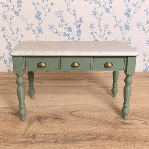 May include: A miniature table with a sage green wooden base and a speckled off-white top. The table features four turned legs and three front-facing drawers, each equipped with a small, semi-circular brass-toned metal pull handle.