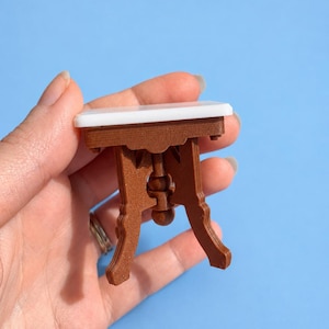 May include: A miniature table with a smooth white rectangular top and a brown base featuring ornate, curved legs and a central vertical support column with a spherical accent. The structure is constructed with visible layered textures.